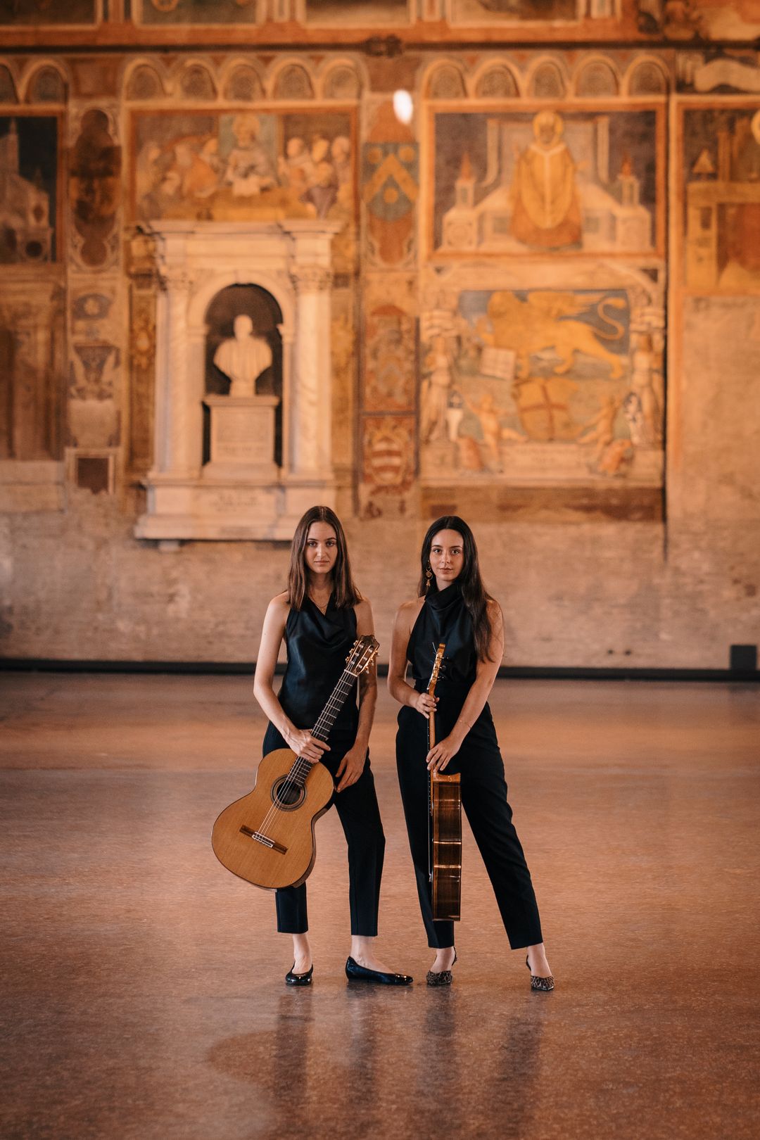 The Daniyah Guitar Duo standing with their guitars in a historic frescoed hall
